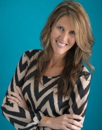 A woman with long, wavy hair smiles and crosses her arms in front of her chest. She is wearing a black and beige zigzag patterned top, embodying the style of The Typical Mom, and standing against a solid turquoise background.