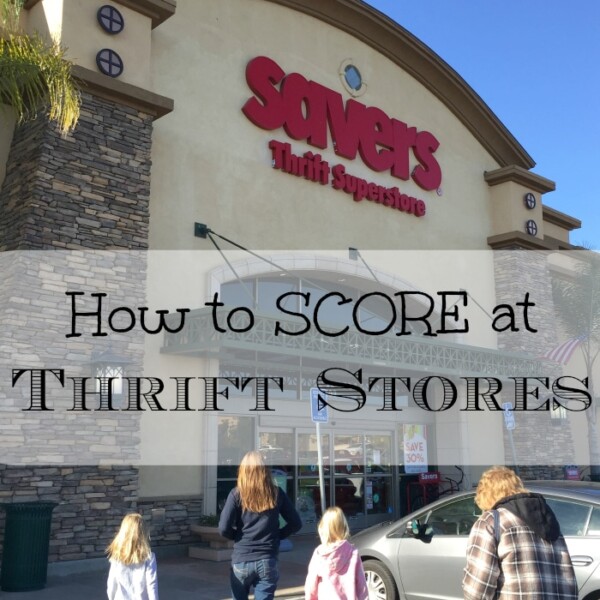 Four people, two adults and two children, walk towards the entrance of a Savers Thrift Superstore. The store has a stone facade and large red sign reading "Savers Thrift Superstore." A sign in front of them reads "How to SCORE at Thrift Stores," offering helpful thrift store shopping tips.