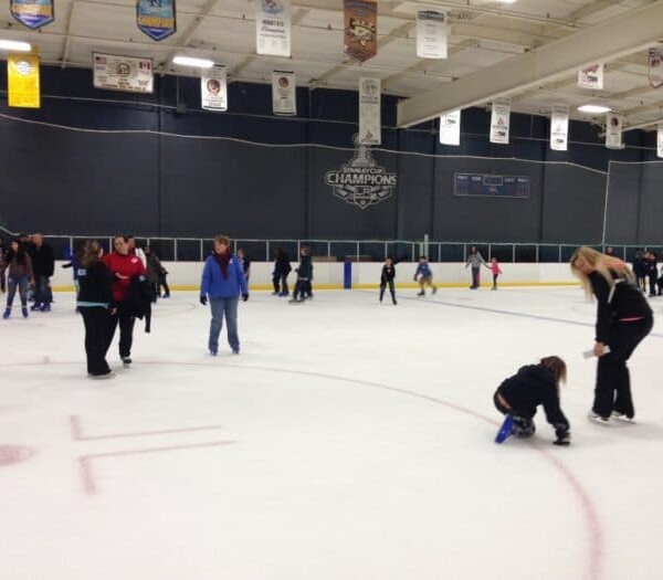 At Icetown Riverside, people glide gracefully across the indoor rink, surrounded by colorful banners. A caring individual kneels to help a child tie their skates while others elegantly skate and socialize around them.