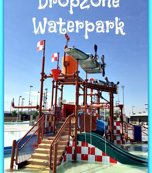 An aquatic play structure with several water slides and climbing features at Perris Water Park's DropZone Waterpark. The structure includes red and white checkered elements and a blue airplane model at the top. The text "DropZone Waterpark" is displayed at the top of the image.