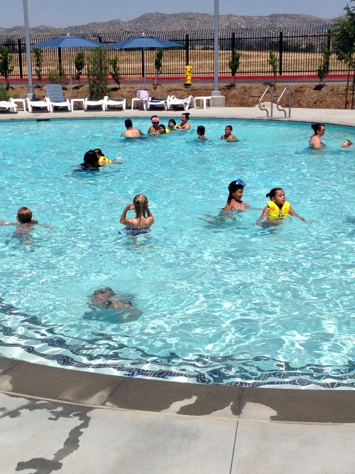 A group of people, including children wearing yellow floatation devices, swim and play in the pool at Perris Water Park. Lounge chairs are aligned along the poolside with a few people sitting and relaxing. A fenced area and a mountainous landscape are visible in the background.