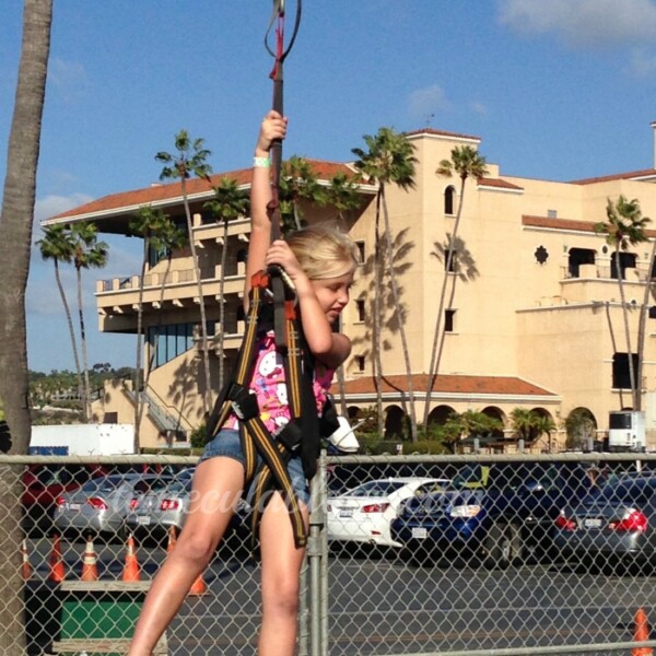 Amid the clear, sunny skies at the San Diego Kids Expo and Fair, a young girl wearing a safety harness climbs a metal pole. She grips a red rope attached to the top while buildings, palm trees, and parked cars in a fenced area provide the lively backdrop.