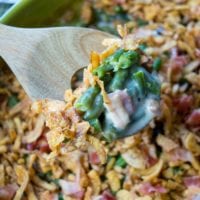 A wooden spoon lifts a serving of easy green bean casserole with crispy onions and diced ham from a green baking dish.