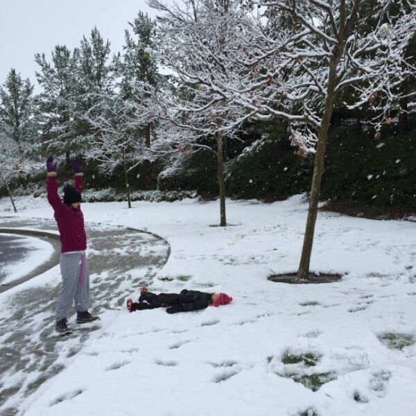 A person stands on a snowy sidewalk with outstretched arms, wearing a pink coat and purple gloves. A second person lies in the snow on their back near a tree. Snow-covered trees and bushes surround the area, creating an enchanting scene reminiscent of snow in Temecula, as flakes continue to fall.