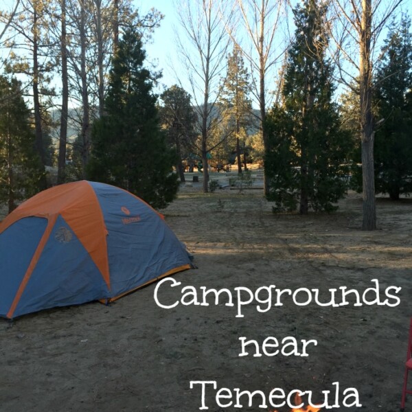 A campsite with a blue and orange tent is set up among tall trees. In the foreground, a campfire is burning in a metal fire ring. The text "Camping near Temecula" is overlayed on the image.