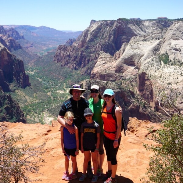 A family of five stands on a rocky cliff at Zion Ponderosa, admiring the scenic canyon view under a clear blue sky.