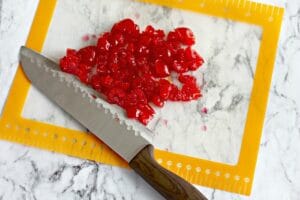 Chopped red candied cherries, perfect for Maraschino Cherry Bread, are spread on a cutting mat with a knife on a marble countertop.