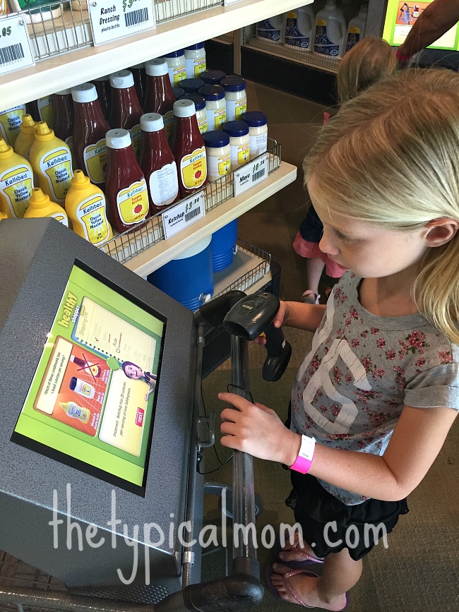 In the aisle brimming with condiments, a girl explores the wonders of modern technology on a touchscreen kiosk. It's like bringing a piece of the Discovery Science Center Santa Ana into her grocery adventure, blending learning with everyday errands.