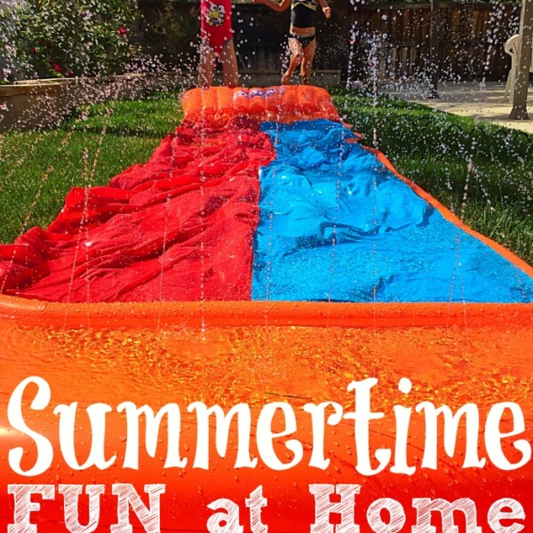 Two children eagerly enjoy summer activities for children on a backyard water slide with sprinkles under a sunny sky. Text overlay reads Summertime Fun at Home.