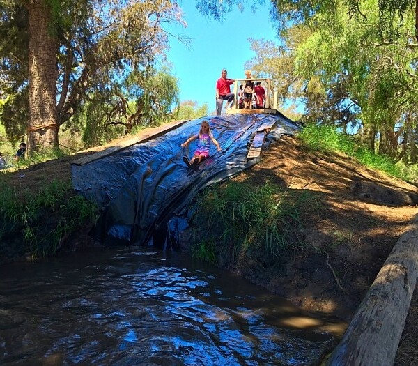 A child joyfully slides down a makeshift tarp-covered slide on a grassy hill, landing near a shimmering body of water. With trees gently swaying in the background, it's reminiscent of simpler times before the thrilling water parks in Southern California became popular.