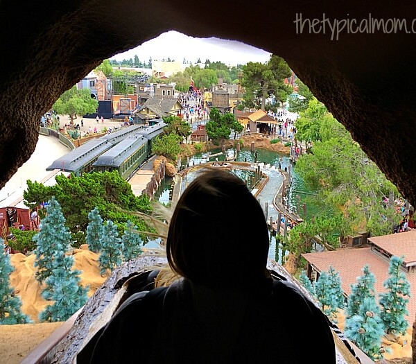 View from inside a tunnel looking out over Knott's Berry Farm, with people, buildings, and trees visible in the distance. A person is silhouetted in the foreground.