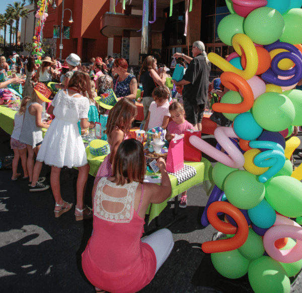 At the kids club promenade in Temecula, a group of people, including children, gather around tables at an outdoor event, surrounded by colorful balloon decorations.