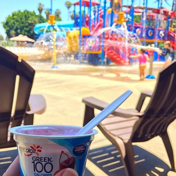 A person enjoys a cup of Yoplait Greek yogurt with a spoon at a water park, seamlessly fitting the vibrant scene. This on-the-go snack pairs perfectly with colorful structures and lounge chairs in the background.