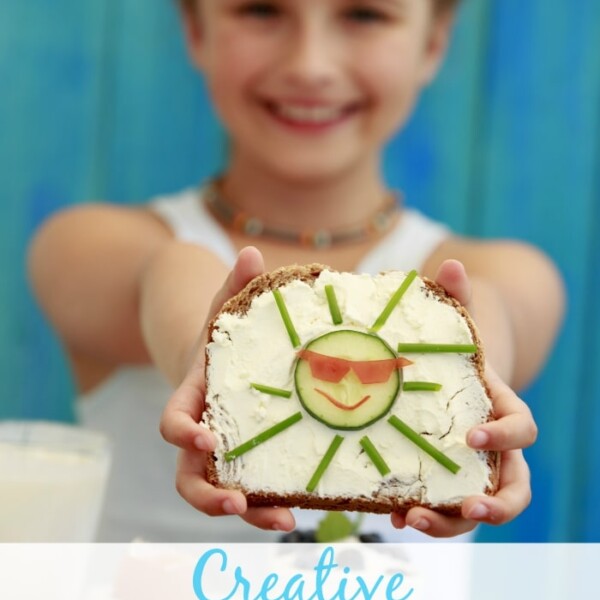 A toddler holds a slice of bread with cream cheese, cucumber, and tomato arranged as a sun face. The text overlay reads Creative School Lunch Ideas for tiny taste buds.