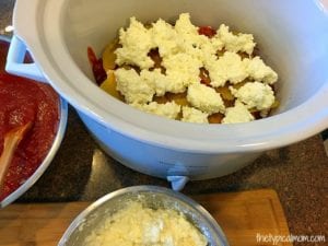 Easy crockpot lasagna recipe featuring uncooked lasagna topped with ricotta cheese in a crockpot. Tomato sauce waits in a nearby bowl with a wooden spoon ready on the side.