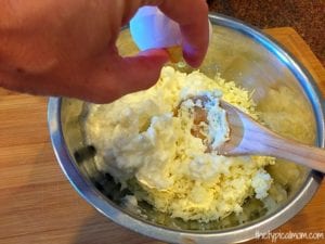 A hand cracks an egg into a metal bowl containing a mixture of shredded cheese and cream, with a wooden spoon in the bowl, preparing the filling for an easy crockpot lasagna recipe.