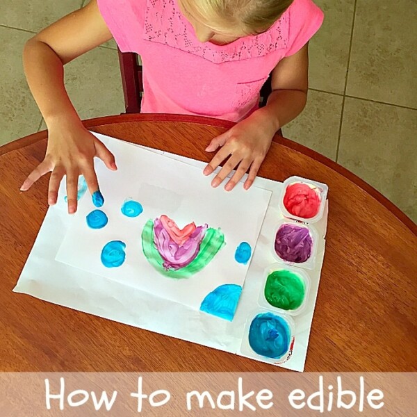 A child joyfully creates vibrant artwork at the table using homemade edible fingerpaint, their tiny fingers exploring various colors from small containers.