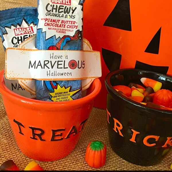 A superhero Halloween-themed setup features Marvel granola bars in an orange "Treat" bowl beside a black "Trick" bowl brimming with Halloween candy, all artfully arranged on a wooden surface.