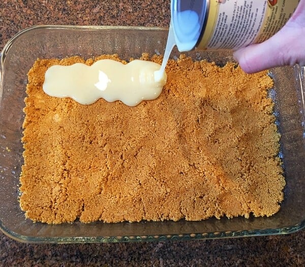 A hand expertly pours condensed milk from a can over a layer of crushed graham crackers in a glass baking dish, setting the foundation for a delectable cookie bar recipe on the speckled countertop.