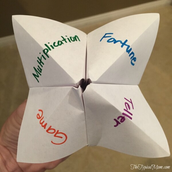 A hand holding a paper fortune teller, cleverly crafted as a multiplication game, features the words Multiplication, Fortune, Game, and Teller inscribed on its flaps.