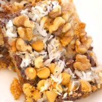 Close-up of cookie bars from a scrumptious recipe, topped with shredded coconut, walnuts, and butterscotch chips. These delectable bars boast a layered texture with chocolate chips and a crumbly base, elegantly placed on a white plate.