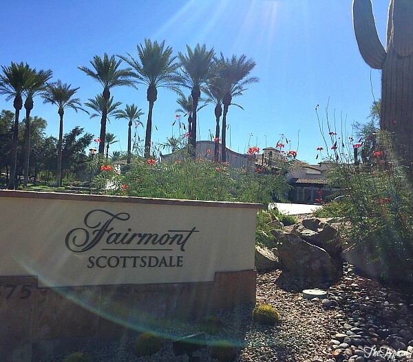 Entrance to the Fairmont Scottsdale Princess, with palm trees, cacti, and vibrant flowers flourishing in the foreground under a clear blue sky.