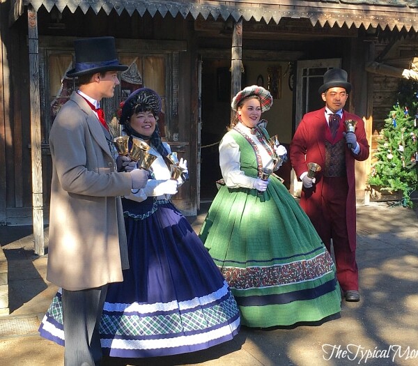 Four people dressed in Victorian-era costumes stand outdoors at Knott's Merry Farm, holding bells in front of a charming wooden building adorned with festive holiday decorations.