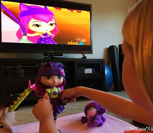 A child plays with a doll resembling a TV character in purple and green attire, capturing the magic of friendship with little charmers. Nearby, a stuffed toy lies on the floor.