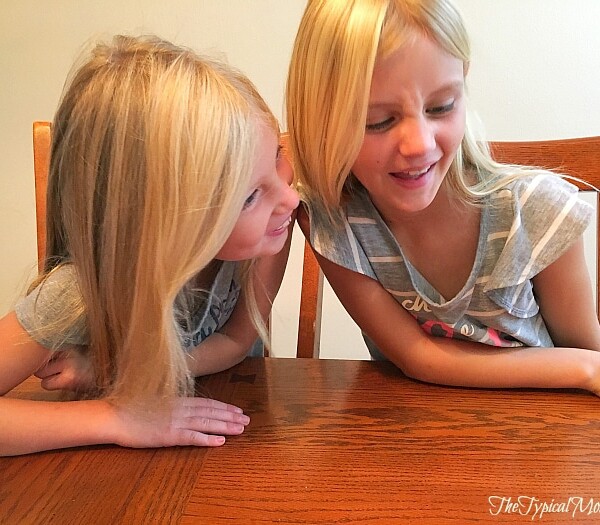 Two young girls with long blonde hair share a moment, leaning over a wooden table during Hasbro family game night. Both wear gray shirts and have expressive faces.