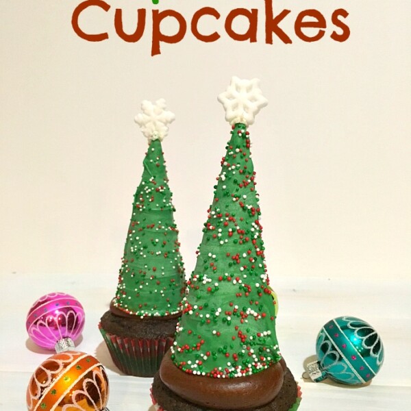 Two cupcakes decorated with green frosting shaped like Christmas trees, topped with star-shaped candies. Colorful sprinkles adorn the trees, while two small decorative ornaments sit beside them. Text reads: Delight in Christmas Tree Cupcakes!