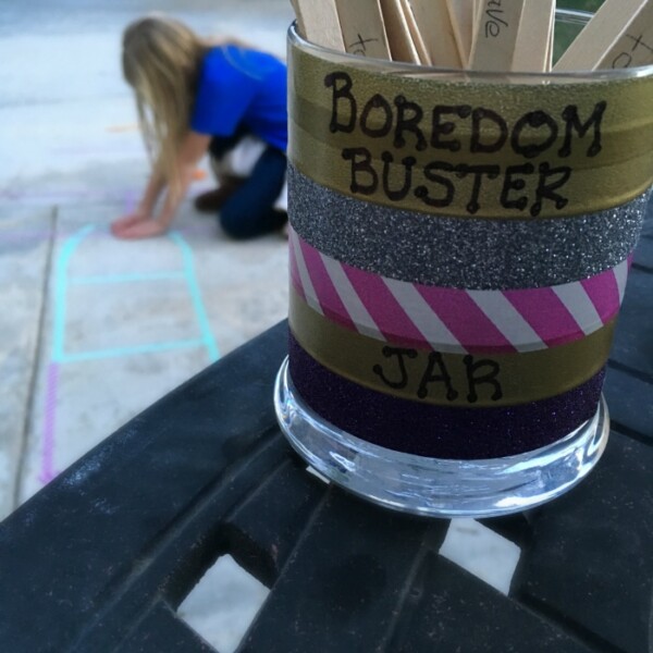 A boredom buster jar filled with wooden sticks sits on a table, while in the background, a child immerses themselves in vibrant chalk art on the pavement.