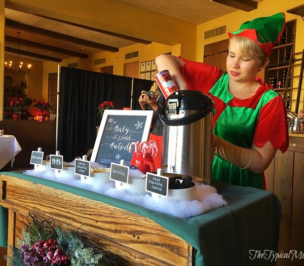 At Downtown Disney, an elf-clad person expertly crafts a beverage at a festive drink station adorned with holiday decorations and a chalkboard sign, capturing the magic reminiscent of Breakfast with Santa.