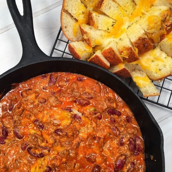 A skillet of cheesy chili with beans sits beside a loaf of pull-apart bread on a cooling rack.