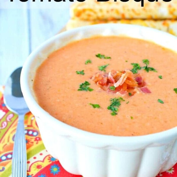 A bowl of creamy tomato bisque, crafted from an easy tomato bisque recipe, is garnished with herbs and sits beside a spoon on a colorful cloth.