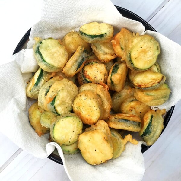 A bowl of salt and pepper zucchini chips rests on paper towels, perfectly crisp on a light wooden surface.