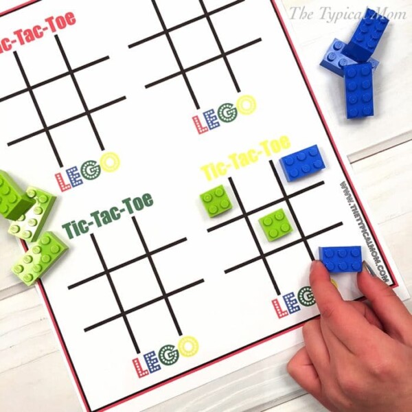 A hand gracefully places blue LEGO bricks onto the tic-tac-toe board, where grids are playfully labeled LEGO Tic-Tac-Toe. Scattered around are green and blue bricks, adding to the vibrant scene set against a soft wood-textured white background.