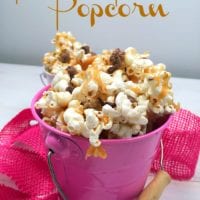 A pink pail brimming with peanut butter chocolate popcorn, sprinkled with delectable chocolate pieces, sits beside a soft pink fabric. Above it, the text reads: Peanut Butter Popcorn.