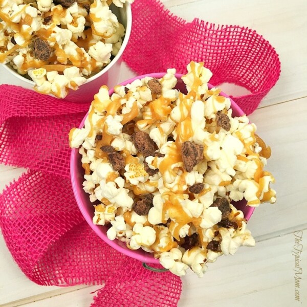 A bowl of peanut butter chocolate popcorn drizzled with caramel, elegantly placed on a pink fabric.