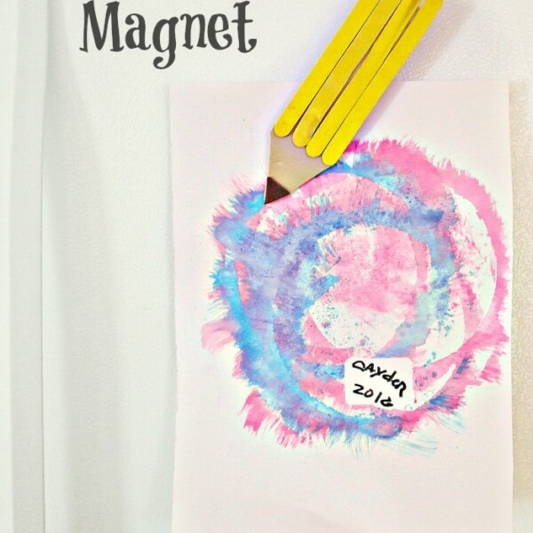 A charming popsicle stick magnet craft holds a painted paper on the refrigerator. The painting features circular blue and pink patterns with a label reading Abby 2012.