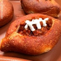 A sloppy joe bread bowl filled with a savory meat and tomato mixture, topped with a zigzag of white dressing resembling football laces, all served on a brown tray.