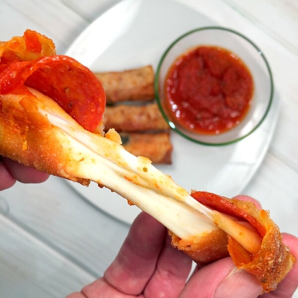 A hand stretches a cheesy pepperoni pizza roll with marinara sauce, surrounded by mouthwatering pizza egg rolls in the background.