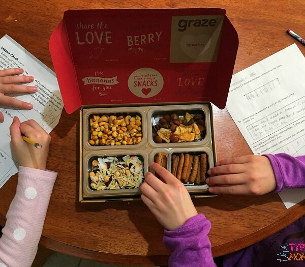 Two children diligently work on their homework at a table, with a snack box filled with assorted nuts and crackers in the center, inspired by a graze promo code that adds an extra crunch to their study session.