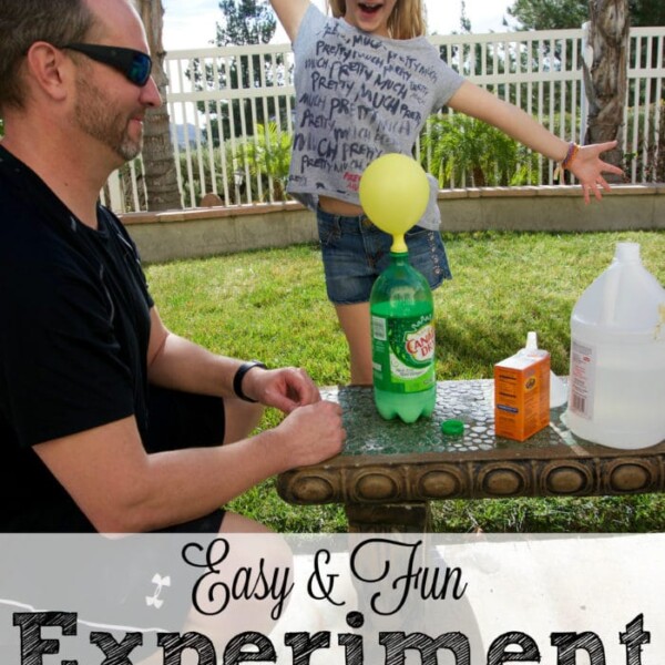 A child with arms raised excitedly stands next to a man, eagerly observing the classic baking soda and vinegar experiment. The setup includes a bottle and balloon, ready to demonstrate the fun science outdoors.