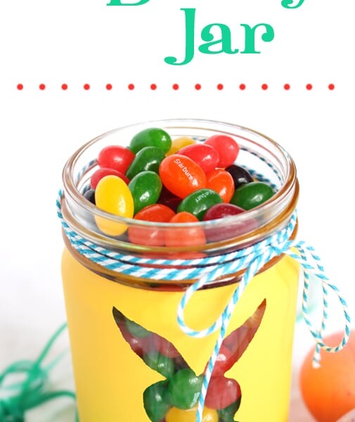 A yellow Easter Bunny jar with a bunny cutout brims with colorful jellybeans, surrounded by decorated eggs and grass-like strands. Text reads DIY Easter Bunny Jar.