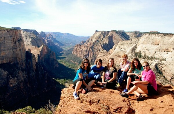 A group of seven women sits on a cliff edge, taking in the vast canyon landscape, during their Zion Ponderosa retreat.