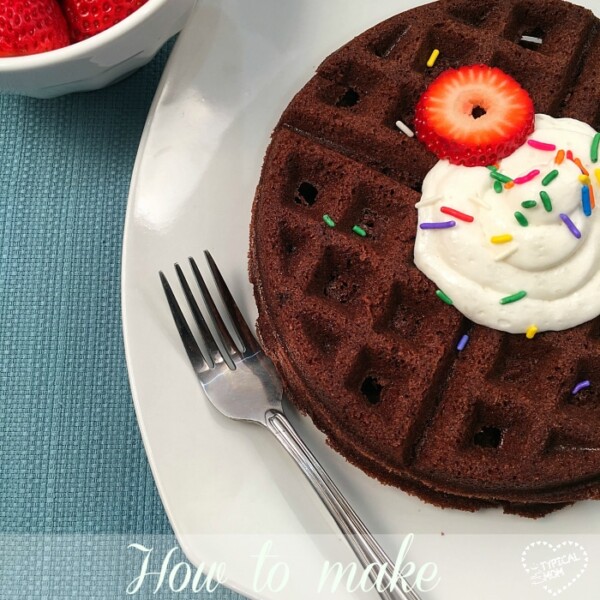 A brownie waffle on a white plate is topped with whipped cream, a strawberry slice, and sprinkles, sitting beside a fork and a small bowl of fresh strawberries.