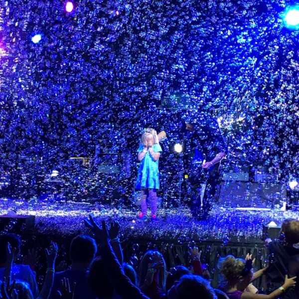 A child in a blue dress stands on stage at Discovery Cube OC's Bubblefest, surrounded by shimmering bubbles and vibrant colored lights, hands outstretched to an audience captivated in the foreground.