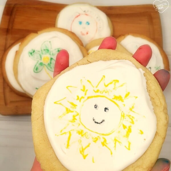 A hand holds a sugar cookie featuring a sun design on white icing, showcasing edible artwork. In the background, other cookies with unique designs rest on a wooden tray, each one illustrating intricate craftsmanship.