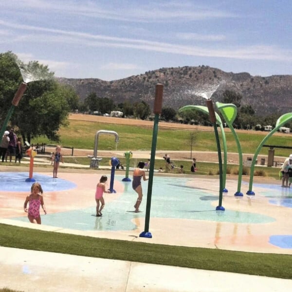 Children frolic in the Lake Skinner splash pad with water features on a sunny day, set against a backdrop of grassy areas and distant hills.