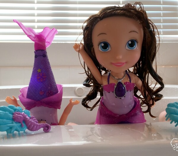 Two bath toys: a mermaid tail sticking out of the water and a doll resembling Sofia the First, both partially submerged in a bathtub, surrounded by plastic sea creatures.